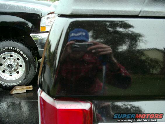 Reflection of person in blue hat taking photo of black SUV rear. Another black vehicle is visible to the left