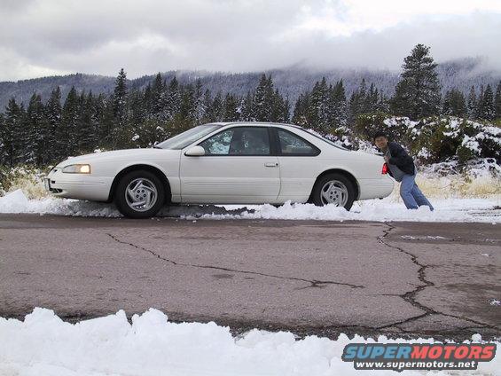 White 1997 Ford Thunderbird stuck in snow, side view. Person pushing