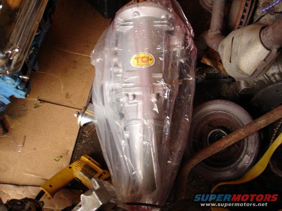 TCI transmission part in plastic wrap with yellow tool and exhaust pipe nearby