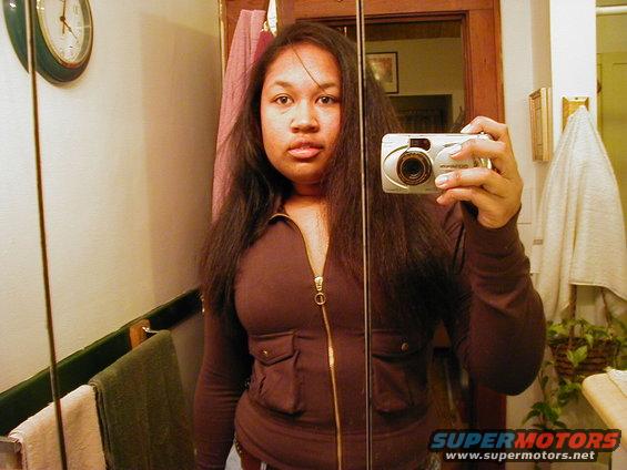 Woman in a brown zip-up jacket takes a selfie in a mirror. A clock is on the wall