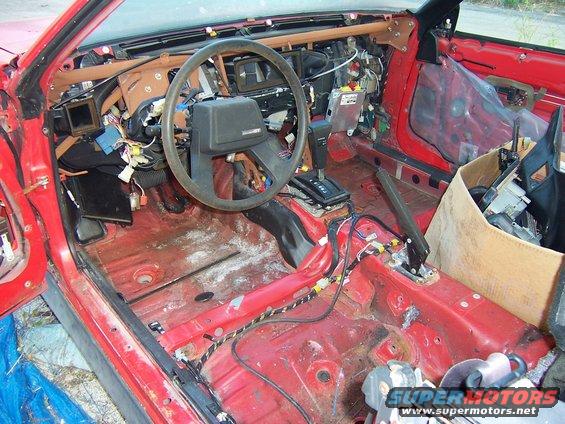 Red car interior with dashboard removed, exposed wiring, and torn seats. A cardboard box filled with parts sits on the passenger side