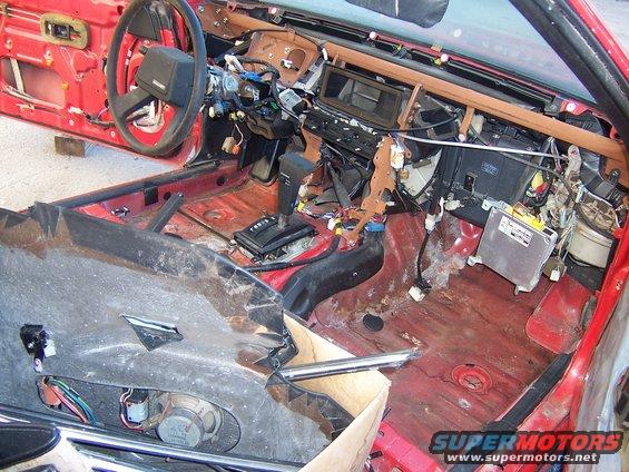 Red car interior stripped down, showing exposed wiring, dashboard components, and floor pan. Automatic transmission shifter visible