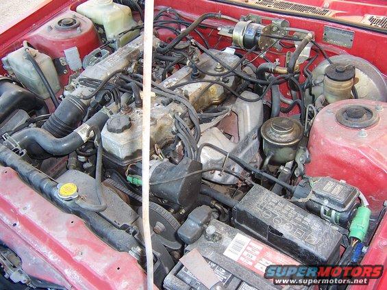 Red 1984 Toyota Celica engine bay with a V6 engine, battery, radiator, and various hoses and wires