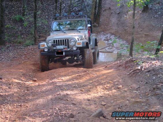Jeep TJ driving on a dirt trail through woods, going through a muddy puddle