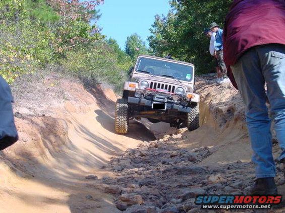 Jeep TJ going down a rocky, rutted dirt trail with trees and people around