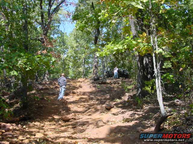 Wooded trail scene with two people and a Jeep in the distance