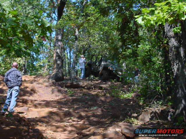 Jeep Wrangler driving up a steep, rocky, wooded trail. Two men stand on the trail watching