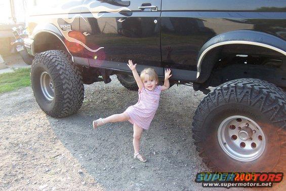 Kid posing under lifted black Ford Bronco with flame decals. Bronco badge visible on fender
