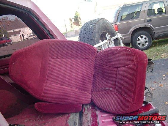Two red velour car seats in the bed of a pickup truck, with a spare tire and a brown SUV in the background