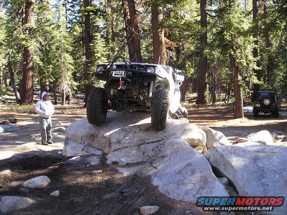 Modified off-road vehicle with large tires on large rocks, a person stands nearby, another vehicle in background