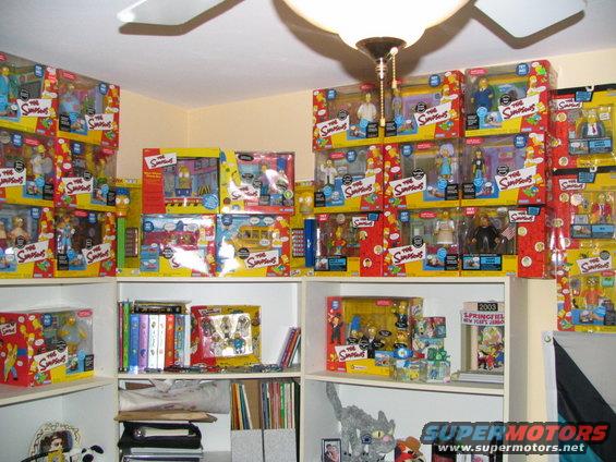 Wall of boxed Simpsons action figures and playsets, with books and a cat statue on shelves below