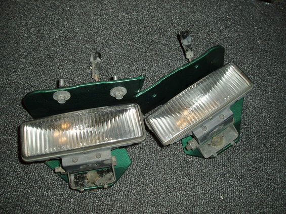 Two green headlight assemblies with clear lenses and metal brackets on a gray carpeted surface