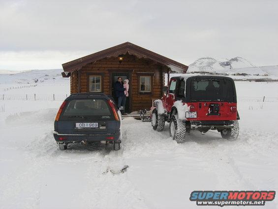 red Jeep Wrangler and blue station wagon parked in snow outside a wooden cabin. Two people and a child stand in the cabin doorway
