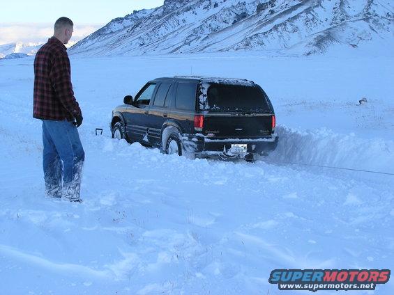 Black SUV stuck in deep snow, a man stands nearby watching a tow rope. Mountains in background
