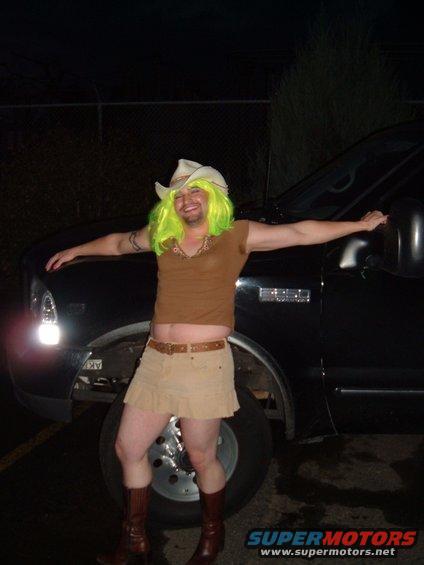 Person in a lime green wig, cowboy hat, brown top, tan skirt, and boots stands in front of a black truck