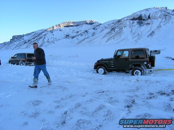Green 2001 Jeep TJ stuck in snow, being towed by a dark SUV