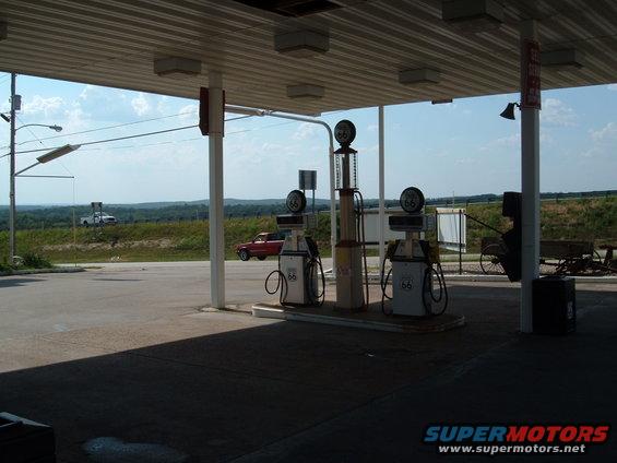 Gas station scene with a red car and a white truck in the distance under a covered canopy with vintage gas pumps