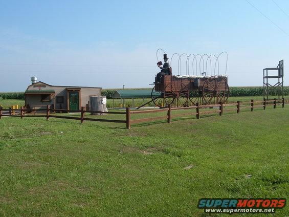 Old wagon with a figure in a top hat, next to a small building and a wooden fence