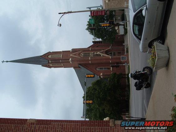 Motorcycle parked on sidewalk in front of a brick church with a tall steeple. A silver car is parked on the street