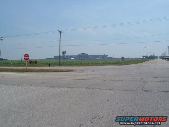 Wide shot of a road intersection with a large, long industrial building in the distance across a field. A 'Do Not Enter' sign is visible