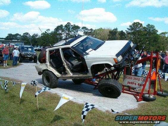 Modified Jeep Cherokee XJ on display with custom suspension and roll cage. Other vehicles and people in background