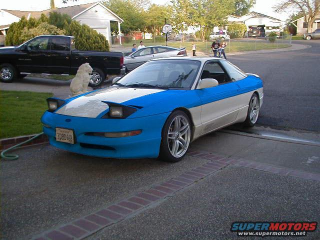 Blue and white 1997 Ford Probe parked in driveway. Black pickup truck and a dog are nearby