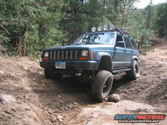 1997 Jeep Cherokee on a steep dirt trail with trees in the background. It has a roof rack with lights