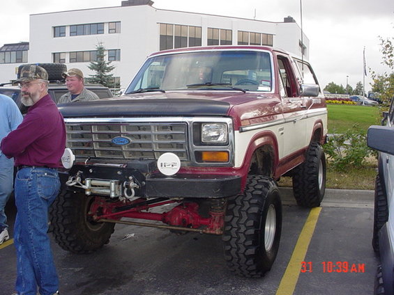 Maroon and white Ford Bronco with aftermarket bumper and winch. Two men stand nearby