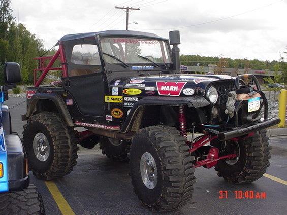 1976 Jeep CJ7 with large off-road tires, snorkel, and numerous stickers. Another blue vehicle is partially visible on the left