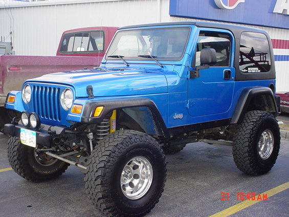 Blue Jeep Wrangler with aftermarket wheels and tires parked next to a red pickup truck