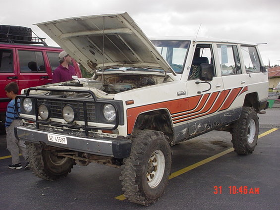 White and orange off-road SUV with hood open, showing engine. Red SUV in background