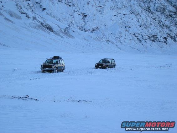 Two SUVs driving in a snowy landscape with mountains in the background