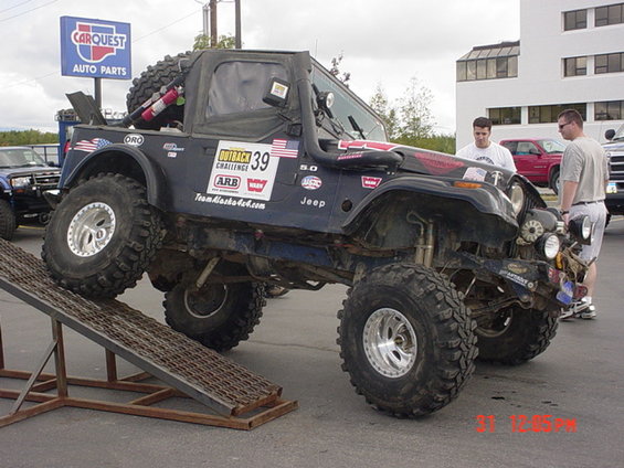 Dark blue Jeep CJ with off-road modifications on a metal ramp, front-right tire elevated