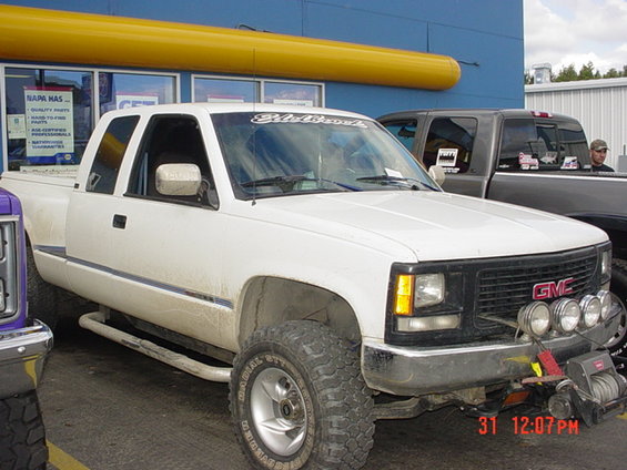 White GMC pickup truck with aftermarket lights and winch, parked in front of a NAPA store