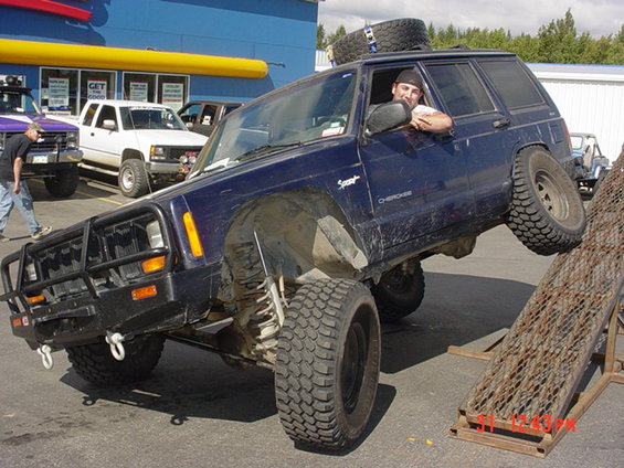 Blue Jeep Cherokee XJ with off-road tires on a ramp, front bull bar, spare tire on roof
