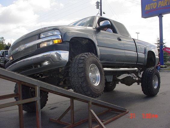 Lifted dark gray pickup truck with large tires on metal ramps, driver visible