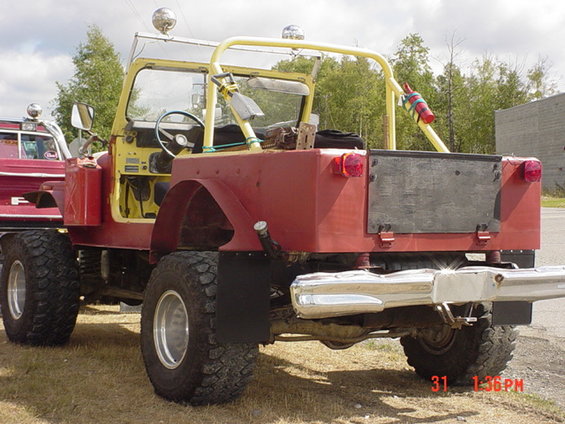 Rear view of a red and yellow modified Jeep with large tires and a roll cage. Another vehicle is partially visible to the left