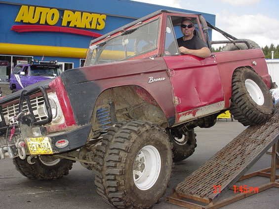 Red Ford Bronco with large tires on a ramp, "Bronco" badge visible. A purple truck is parked behind it. License plate BEX 984