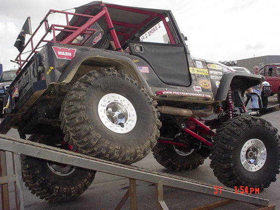 Modified black Jeep Wrangler with large off-road tires on a ramp, red roll cage, and various stickers