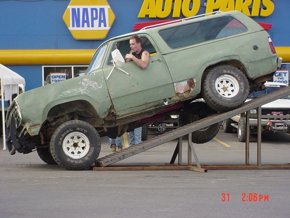 Green SUV with rust spots on a metal ramp, driver looking out window. NAPA Auto Parts store in background