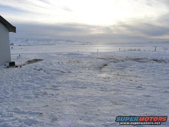 Snowy landscape with a building on the left and mountains in the distance under a cloudy sky
