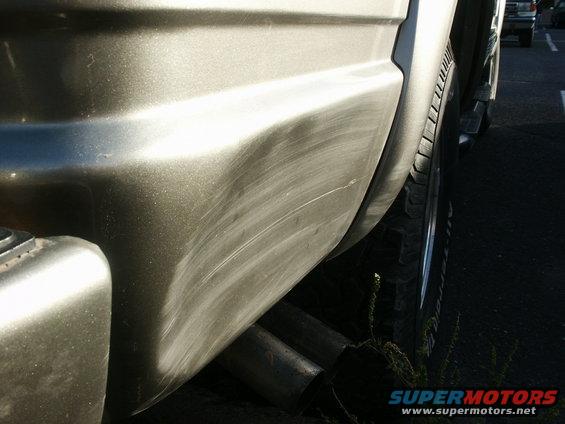 Close-up of a vehicle's rear quarter panel with visible scratches and dual exhaust pipes. Another vehicle is visible in the background