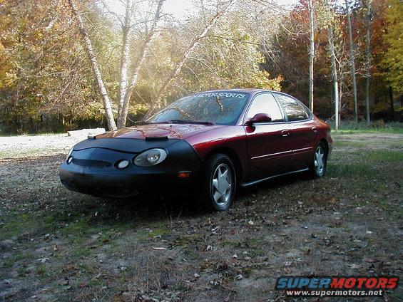 Maroon sedan with a black car bra on the front, parked in a grassy area with trees in the background