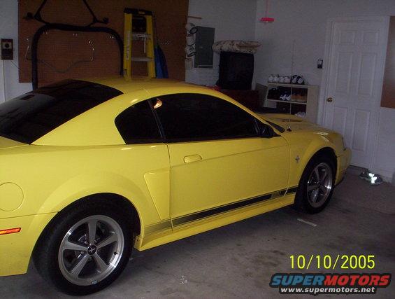 Yellow 2003 Ford Mustang coupe with black racing stripes on the side, parked in a garage
