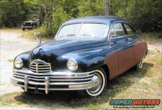 Dark blue sedan with rusty fender, front 3/4 view. Another vehicle in background