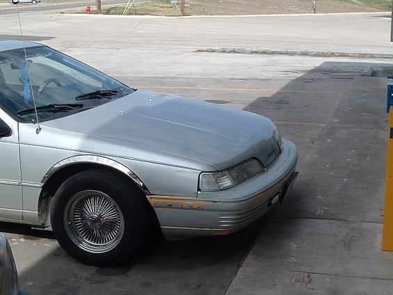 Silver sedan with wire wheels parked on concrete. Front fender and bumper show some damage