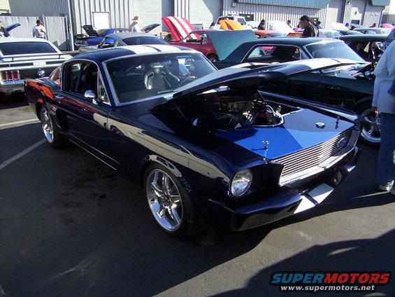 Dark blue classic Ford Mustang with white racing stripes, hood open showing engine. Other cars visible in background