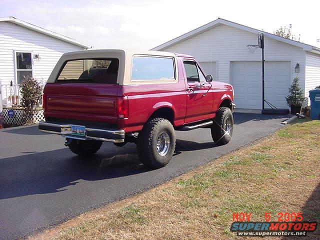 Red Ford Bronco parked in driveway, tan top, lifted suspension, large tires. Garage in background