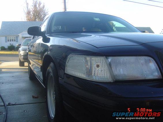 Dark sedan, front quarter view, showing headlights and grille. Another car is parked behind it