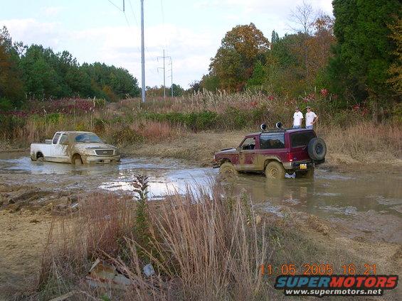 Maroon Ford Bronco II and white Ford pickup truck stuck in mud, with two people standing on Bronco II